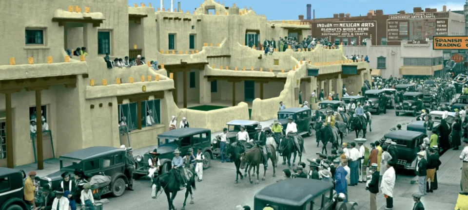A historic photograph captures a busy downtown parade with vintage automobiles and spectators lining the Pueblo-style.