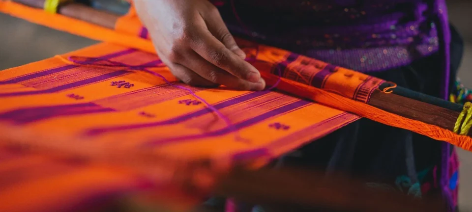 A person's hand guides purple thread into a pattern on vibrant orange fabric stretched across a loom.