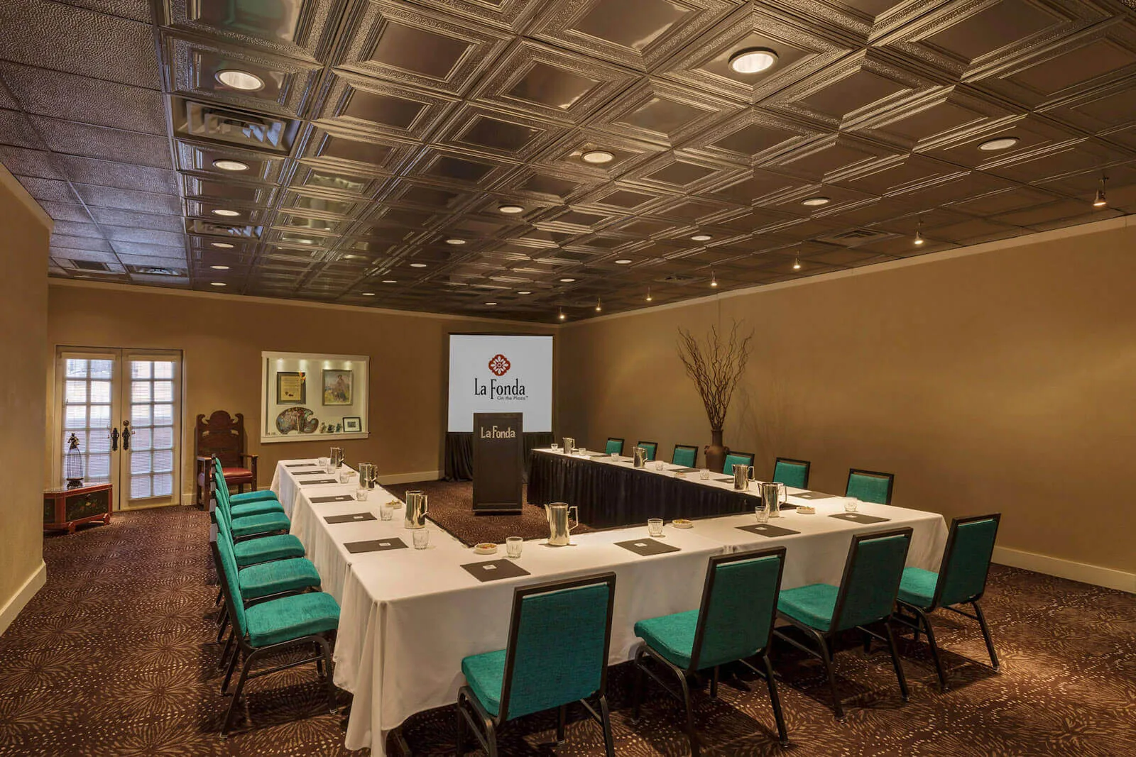 This elegant event space features a U-shaped table setup, teal chairs, and a decorative pressed tin ceiling.