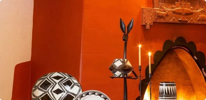 Native American patterned pottery and burning candles sit beside an illuminated adobe fireplace set into an orange wall.