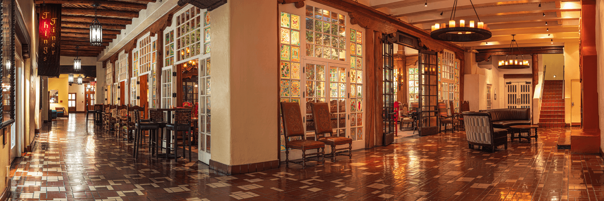 Welcome to La Fonda on the Plaza, where a spacious, warmly-lit hallway with wooden beams and a tiled floor invites you in. A cozy seating area with chairs and a sofa offers comfort, while decorative glass panels add elegance. Overhead, chandeliers sparkle as stairs beckon in the background.