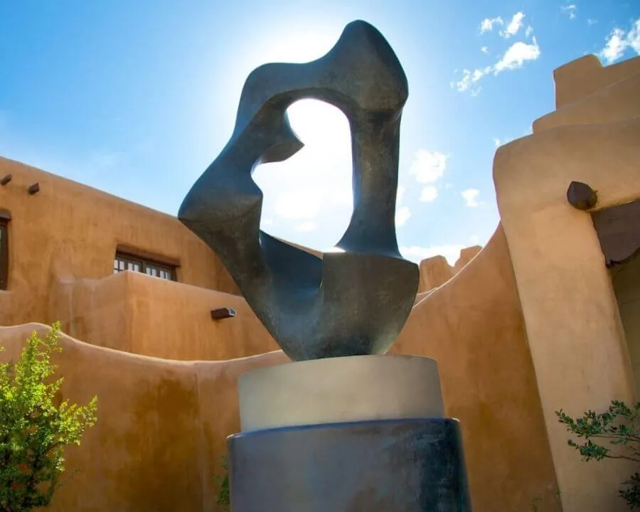 A striking abstract bronze sculpture stands outside against the warm, curving adobe exterior walls under a bright blue.