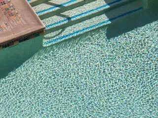 The crystal clear water of the swimming pool reflects sunlight onto the blue-tiled entry steps.