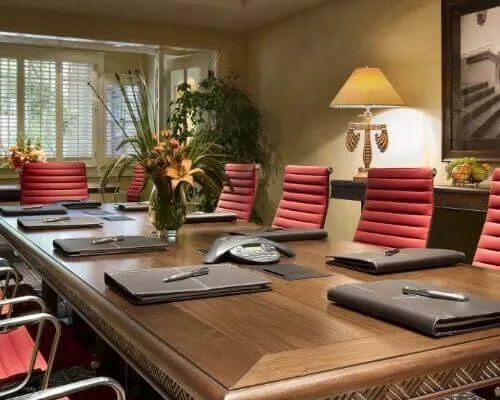 A professional boardroom setup in a small event space with a wooden table, red chairs, and natural light.