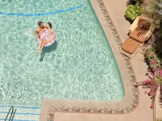 A couple is relaxing on a float in the sunlit outdoor swimming pool surrounded by a tiled deck.