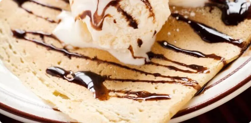 A folded crepe dessert is generously drizzled with dark chocolate sauce and topped with a large scoop of vanilla ice.