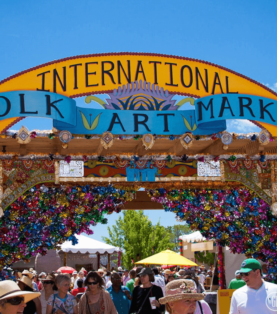 A vibrant archway decorated with colorful flowers and patterns marks the entrance to the International Folk Art Market, welcoming visitors akin to the charm of La Fonda on the Plaza. Crowds of people wearing hats and summer clothing walk underneath on a sunny day.