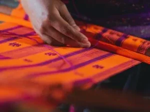 A hand carefully works a loom, weaving a vibrant pattern of orange and purple threads into a finished textile.