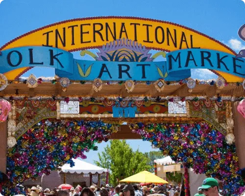 A vibrant, festive archway decorated with ribbons marks the entrance to a crowded international folk art market.
