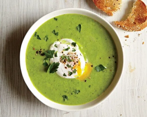 A vibrant green soup is garnished with a poached egg and herbs, alongside toasted bread.