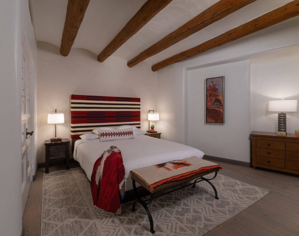 A comfortable santa fe hotel room displays a large bed, colorful striped headboard, and rustic wooden furniture.