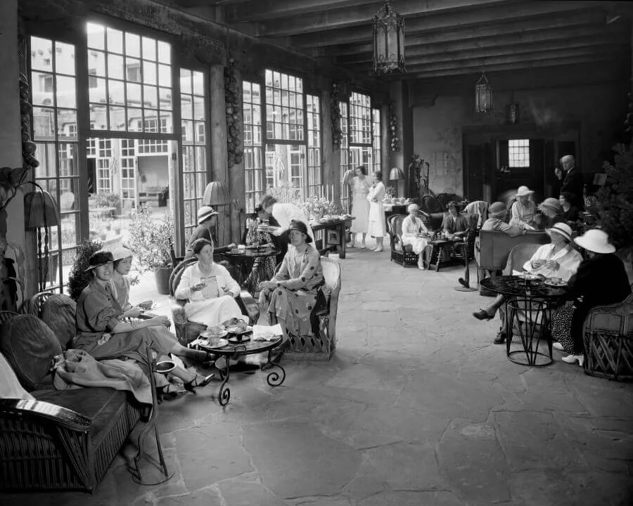 Guests from the 1920s socialize and have tea inside a historic hotel event space with floor-to-ceiling windows.