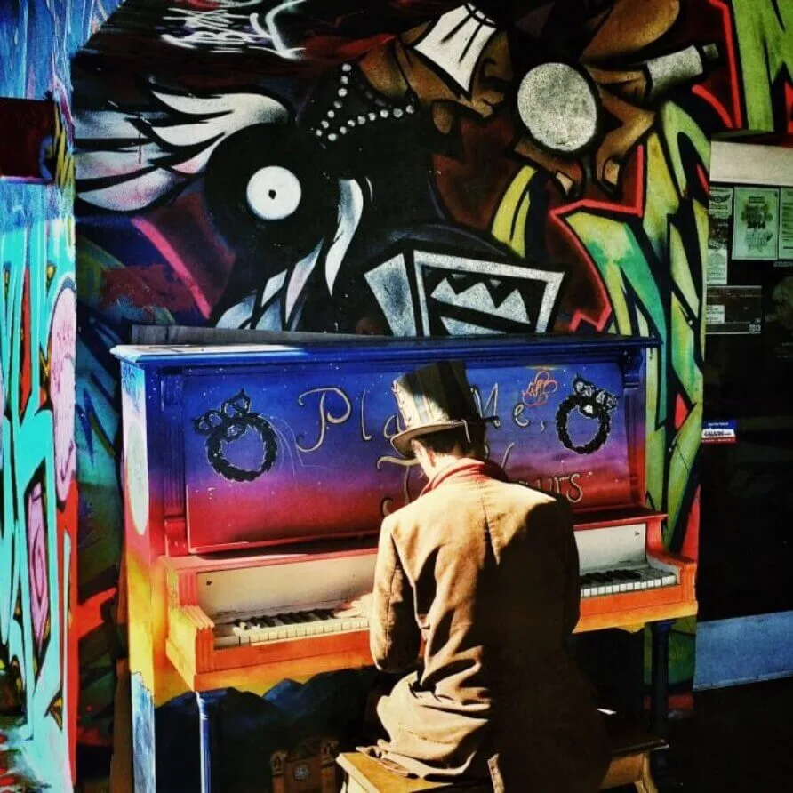 A performer wearing a top hat plays a brightly graffiti-painted piano, highlighting the vibrant scene and live music.