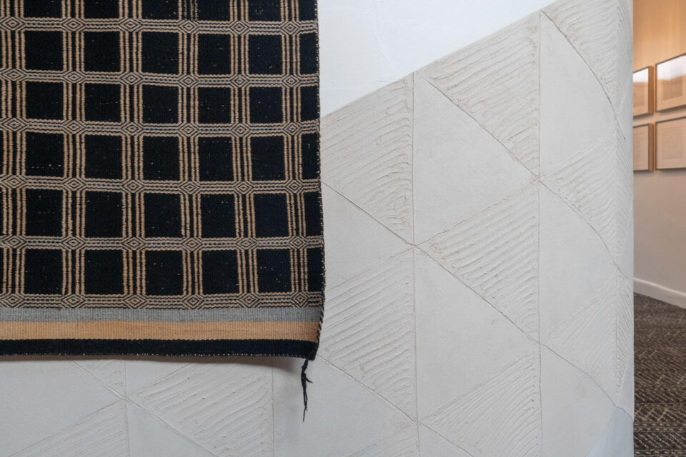 A geometric black and tan woven textile hangs against a curved white textured wall pillar.