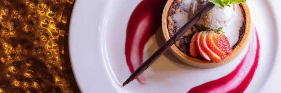 A beautiful dessert tart featuring vanilla ice cream, sliced strawberry, and chocolate is served on a white plate with.