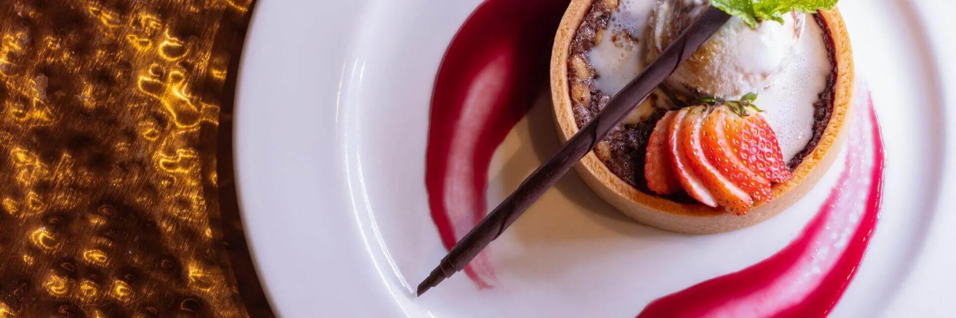 A beautiful dessert tart featuring vanilla ice cream, sliced strawberry, and chocolate is served on a white plate with.