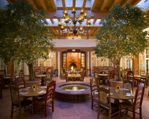 Welcome to La Fonda on the Plaza, a cozy restaurant where a central fountain is surrounded by round tables and chairs. Two large indoor trees beautifully flank the dining area, while a warm chandelier hangs from the wooden beam ceiling, enhancing the inviting atmosphere.