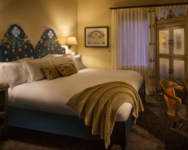 Welcome to La Fonda on the Plaza, where a cozy bedroom awaits with a large bed featuring ornate blue headboards and fluffy pillows. A warm brown throw blanket is draped over the bed, complemented by a wooden cabinet, patterned curtains, and a small lamp for an inviting atmosphere.