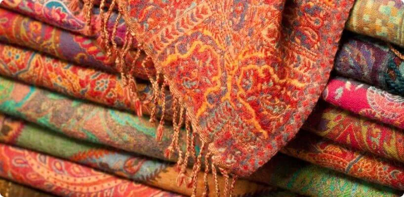 A stacked collection of folded, richly colored artisan shawls with intricate designs and tassels is shown.