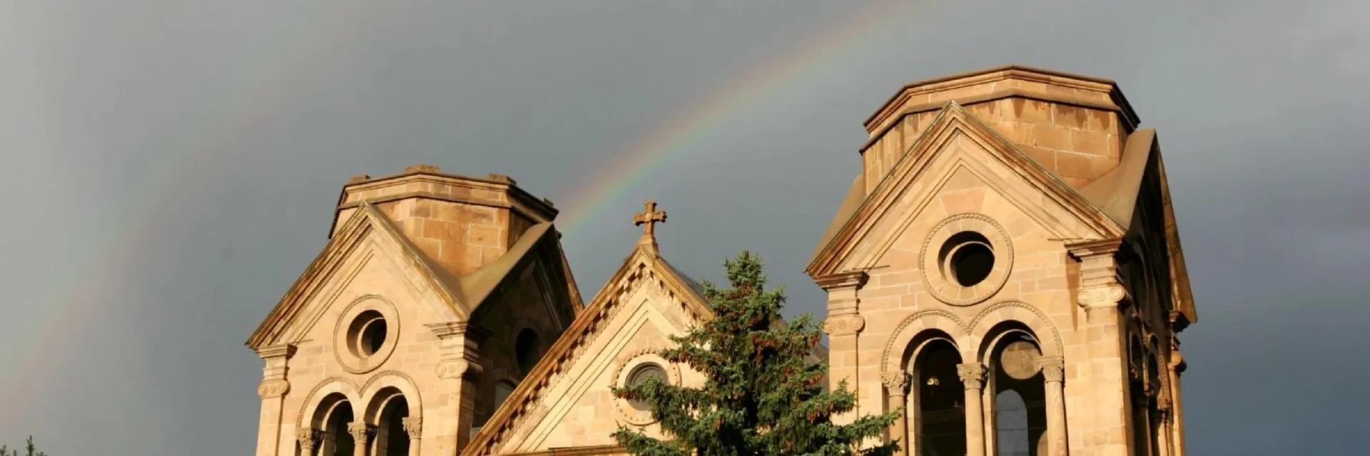A vivid rainbow arches over the historic stone facade and twin towers of the cathedral beneath a dark sky.