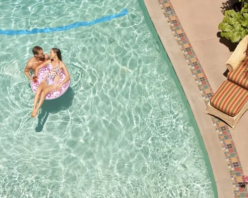 A couple floats on an inflatable ring in the clear water of the outdoor swimming pool next to a striped lounge chair.