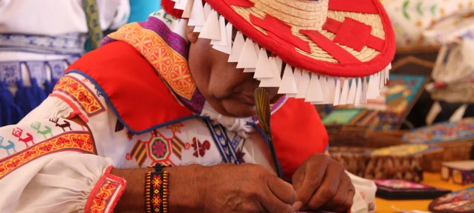 A craftsperson in colorful traditional clothing and a woven hat meticulously works on a detail with a feather.