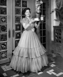 An archival photo shows an attendant in a formal, tiered gown holding a silver service tray inside a richly decorated.
