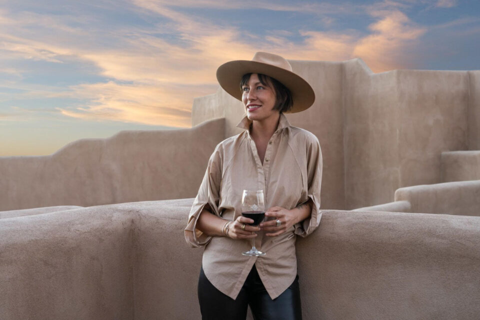 A stylish woman holding red wine relaxes on the adobe-style rooftop terrace overlooking the sunset.