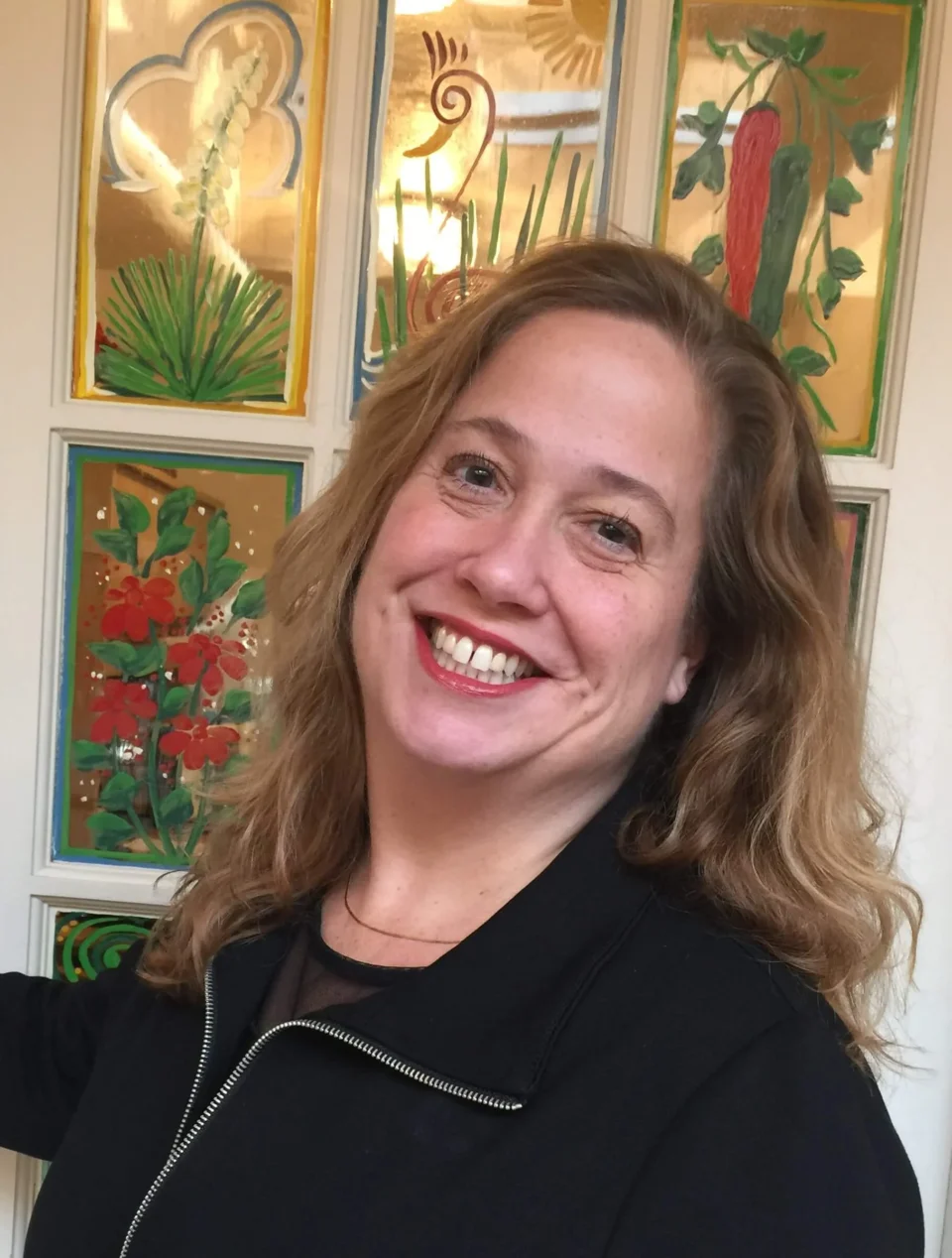 A friendly woman smiles widely in front of artistic panels painted with Southwestern chili peppers and flowers.