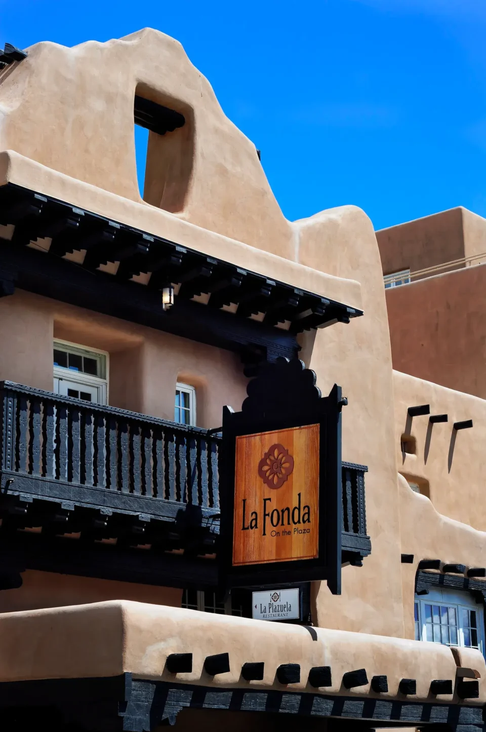 The adobe exterior of La Fonda On The Plaza features a dark wooden balcony and signage for La Plazuela Restaurant.