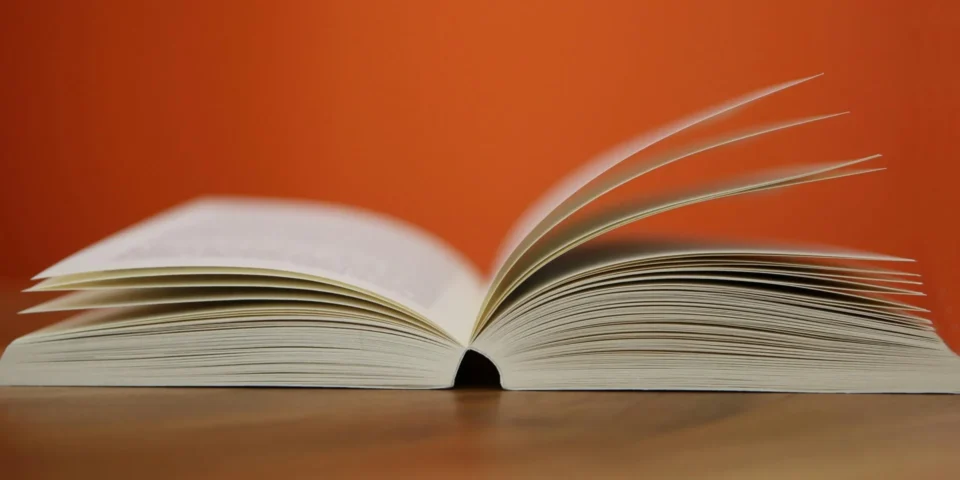 A thick, open book with white pages is displayed on a wooden surface against an orange wall.