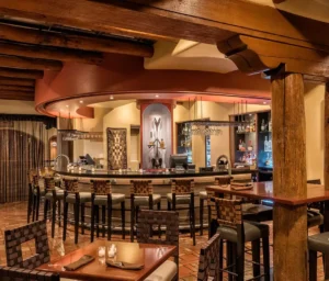 The interior view of the adobe-style bar shows a curved counter, exposed wooden beams, and intimate table seating.