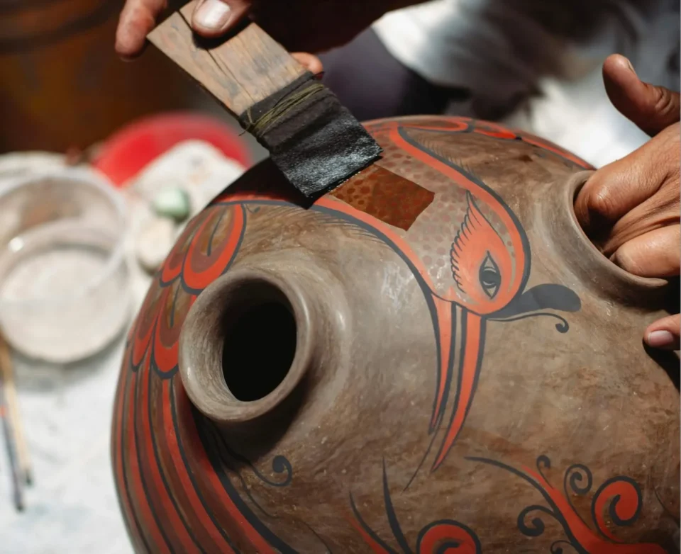 A craftsman applies a finishing layer of glaze to a clay pot featuring vibrant red and black artistic designs.