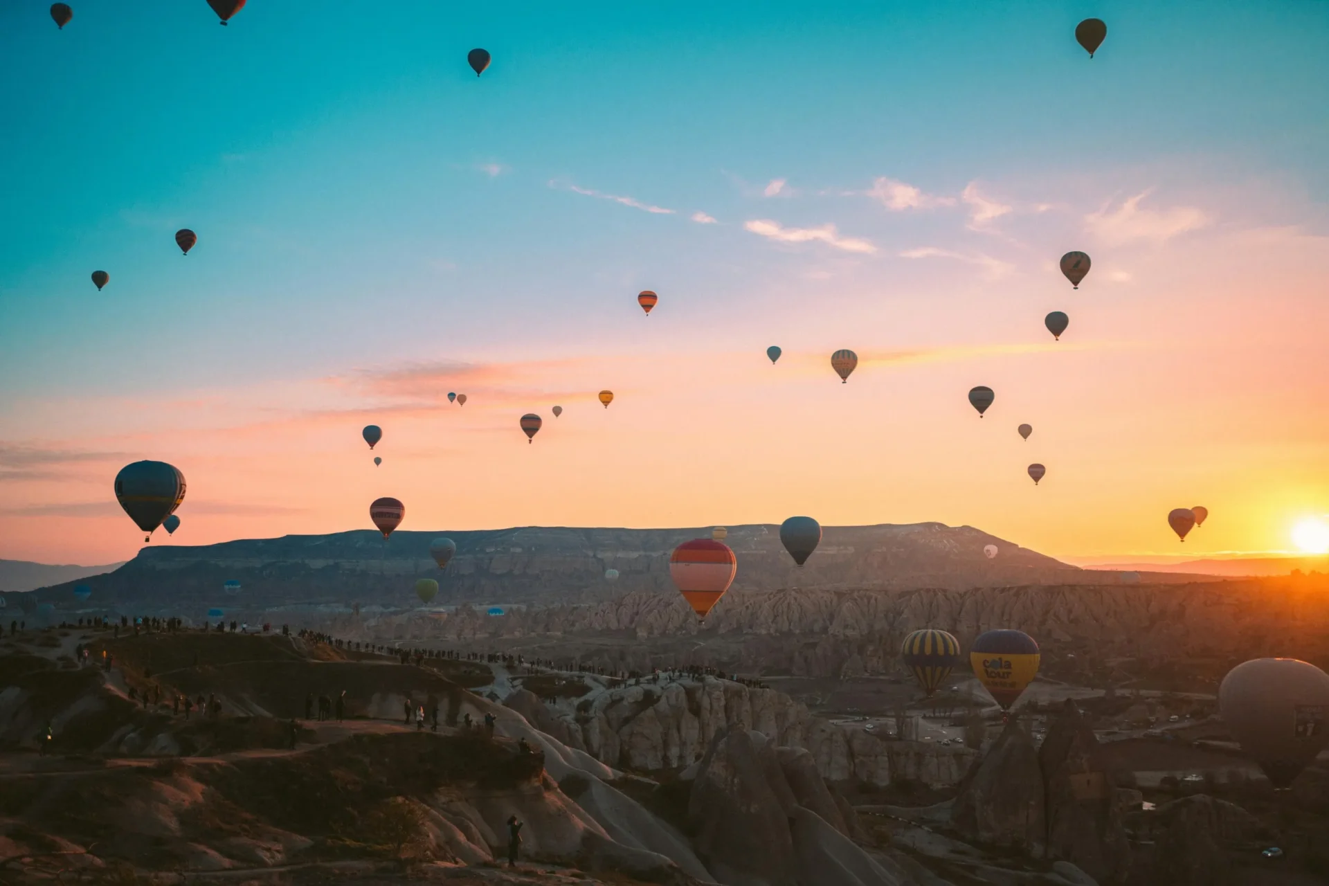 Dozens of colorful hot air balloons float over a rugged valley landscape against a dramatic pink and blue sunrise.