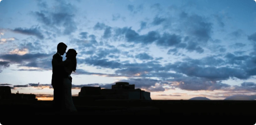 A romantic silhouette of a bride and groom embracing against a beautiful sunset sky above an event space.