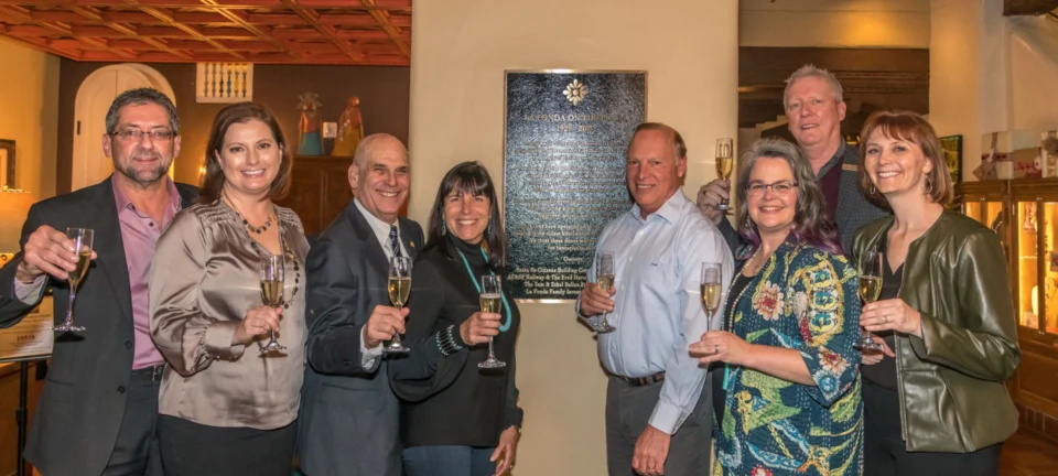 A joyful group of eight guests holds champagne flutes near a commemorative plaque inside the hotel.