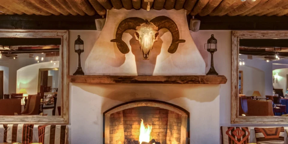A rustic adobe fireplace with a burning fire is centered below a ram's skull mount in a warm, inviting lounge.