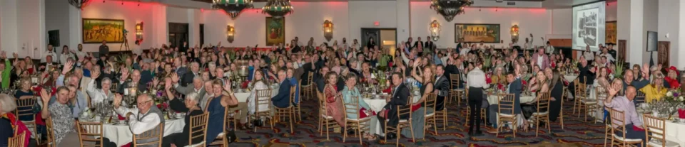 Hundreds of smiling guests are seated at round tables inside the ornate hotel banquet hall.