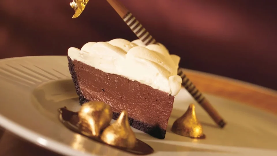 A rich slice of chocolate mousse pie topped with whipped cream and edible gold leaf is elegantly served.