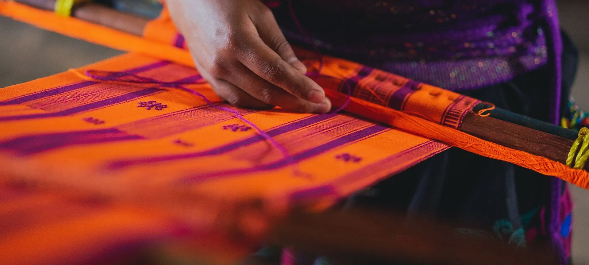 A person's hand guides purple thread into a pattern on vibrant orange fabric stretched across a loom.