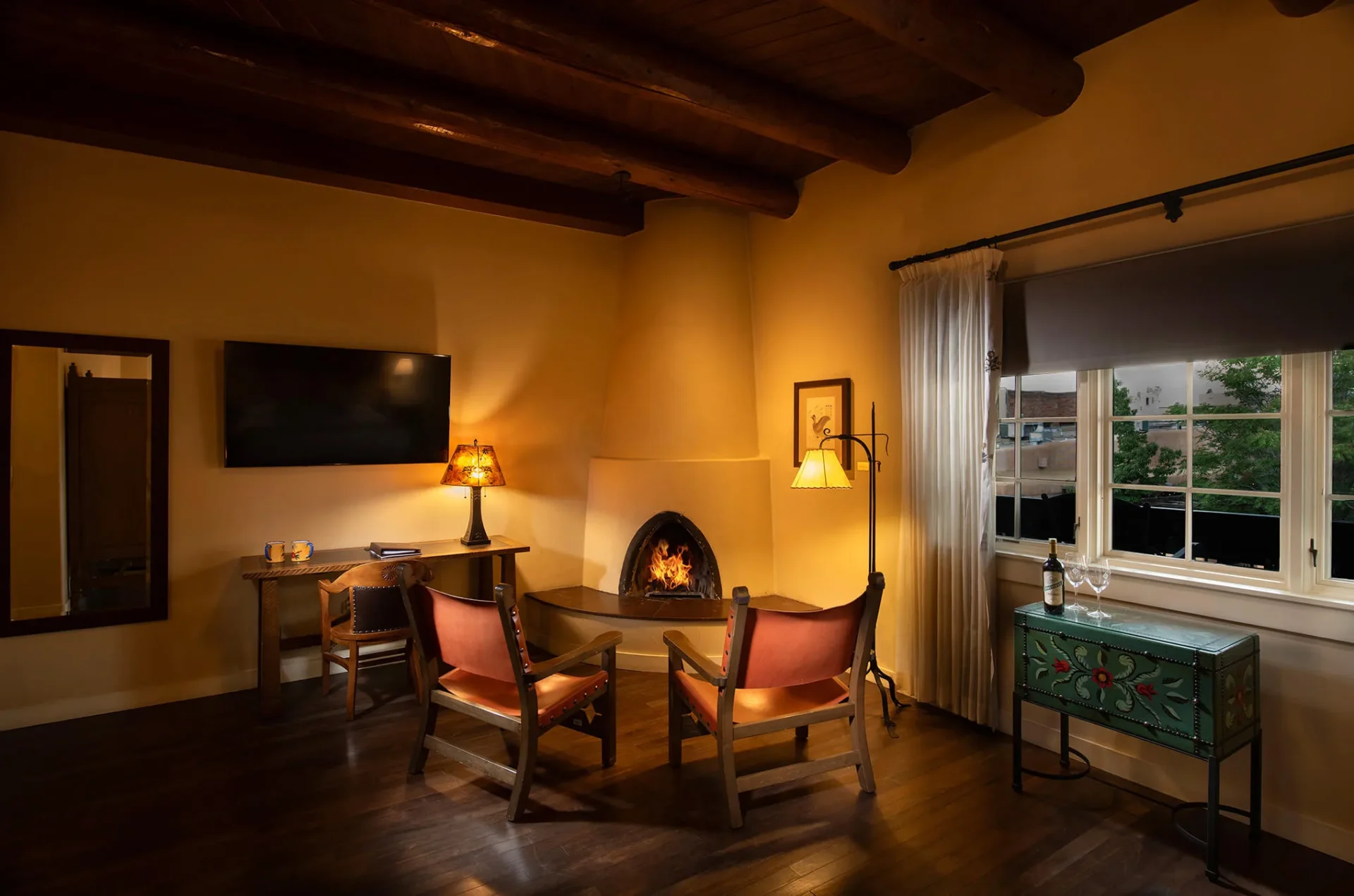 An inviting hotel suite features comfortable seating by a kiva fireplace and exposed wooden ceiling beams.