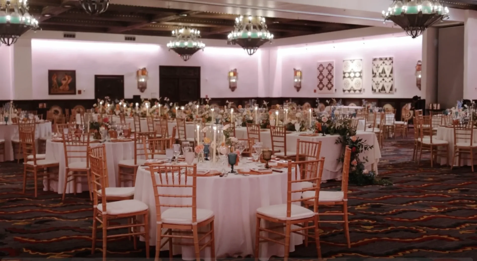 A large banquet hall with round tables set for dining, adorned with white tablecloths, gold chairs, floral arrangements, and candles.