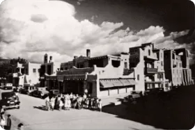 A black and white historical photo captures a busy street with traditional Pueblo architecture and a crowd crossing the.