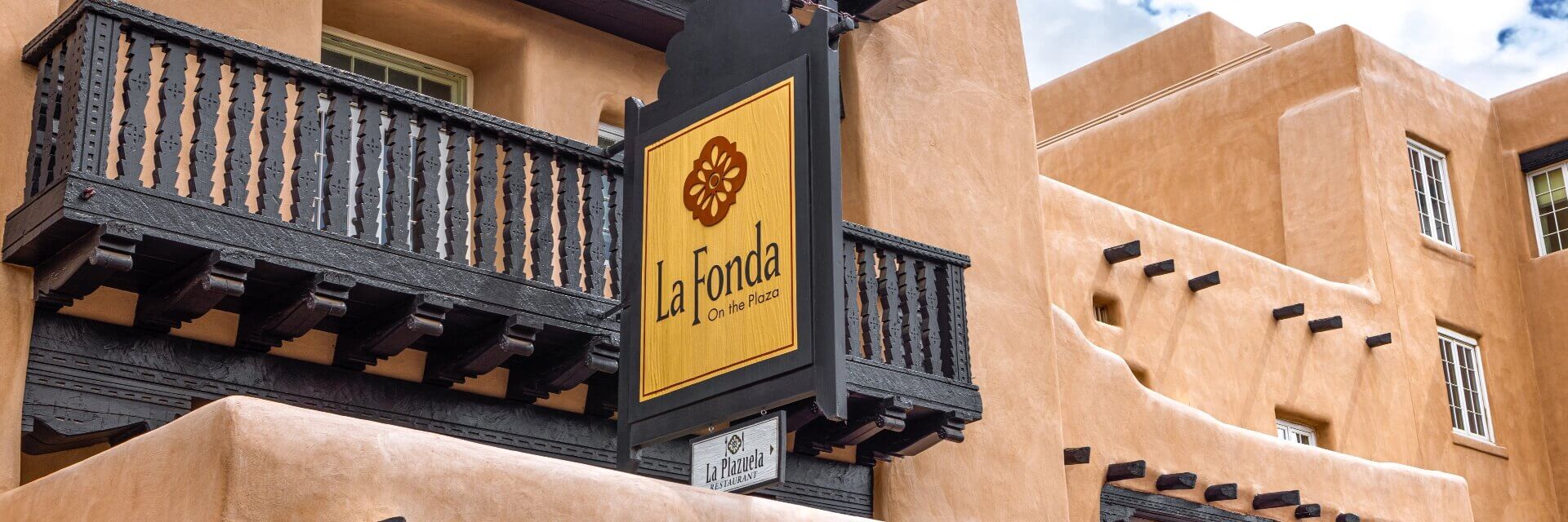 The adobe building exterior displays a dark wooden balcony and the prominent sign for la Fonda On The Plaza.