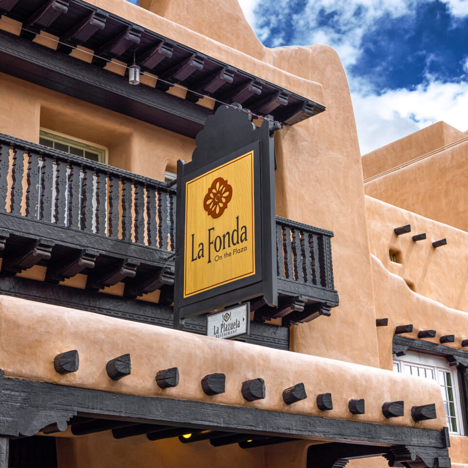The iconic La Fonda On the Plaza sign hangs above the dark wood balcony and the La Plazuela Restaurant entrance.