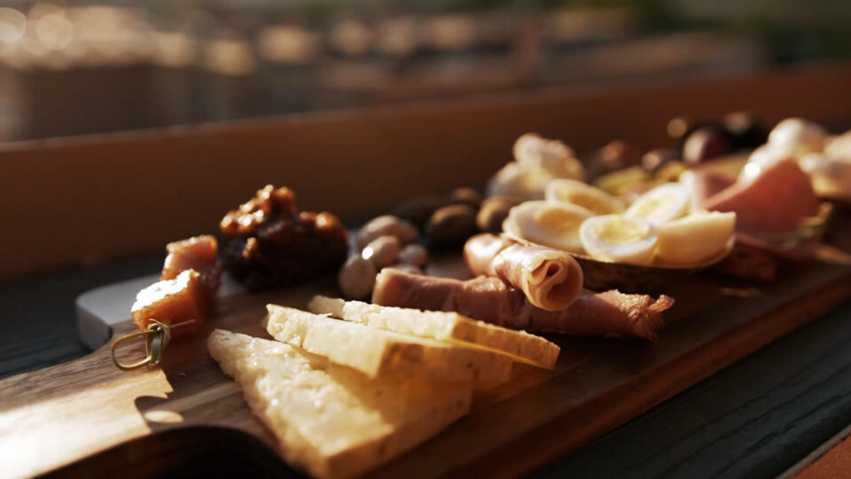 A close-up shows a wooden board topped with hard cheese, rolled prosciutto, eggs, and fruit preserves.