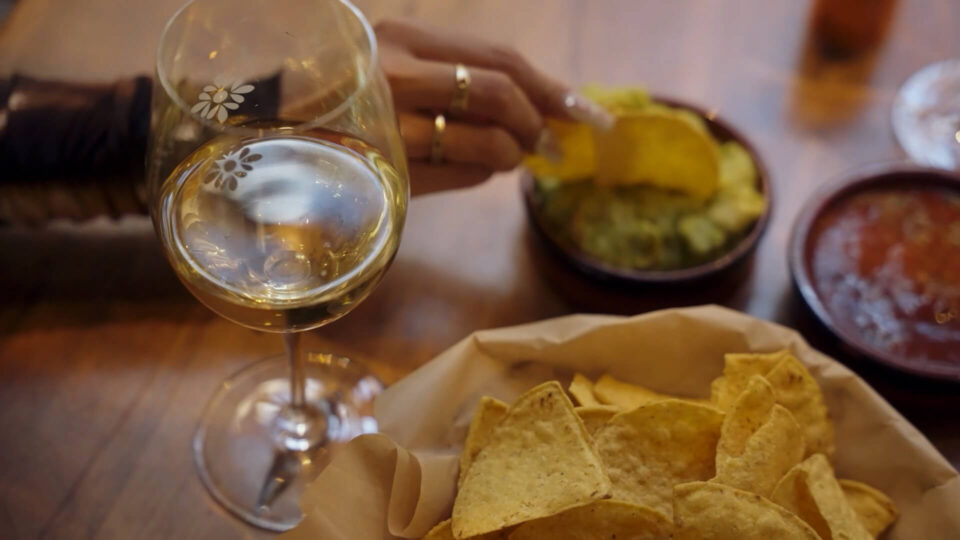 A hand dips a chip into a bowl of guacamole next to a glass of white wine and a bowl of salsa.