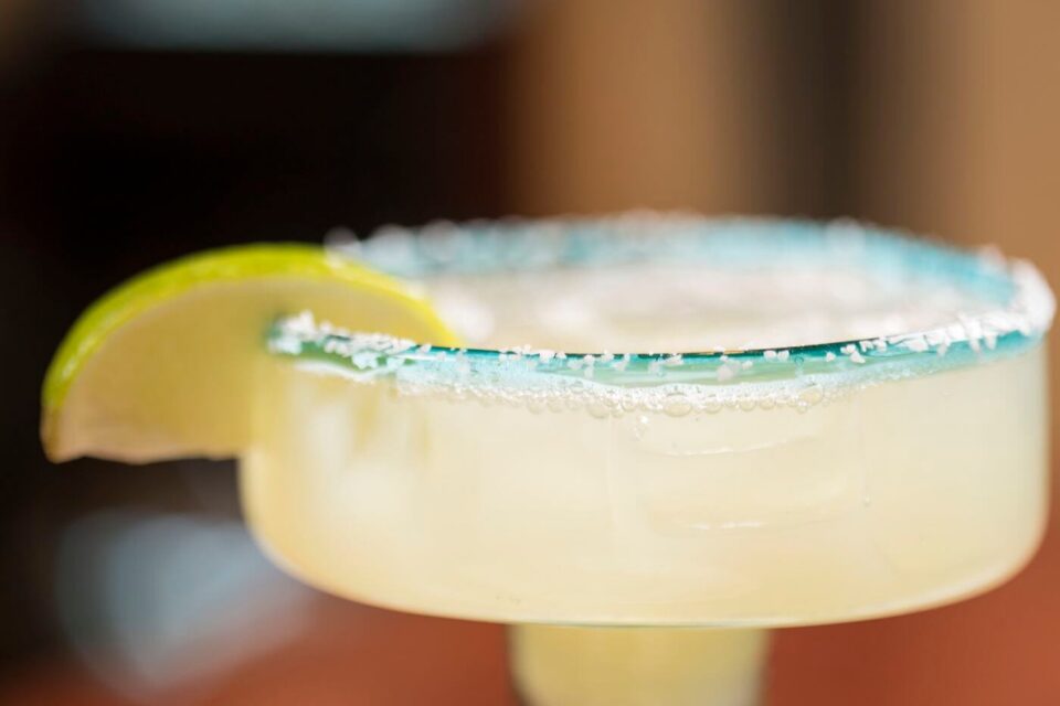 A refreshing margarita cocktail is served in a blue-rimmed, salted glass, garnished with a bright green lime wedge.