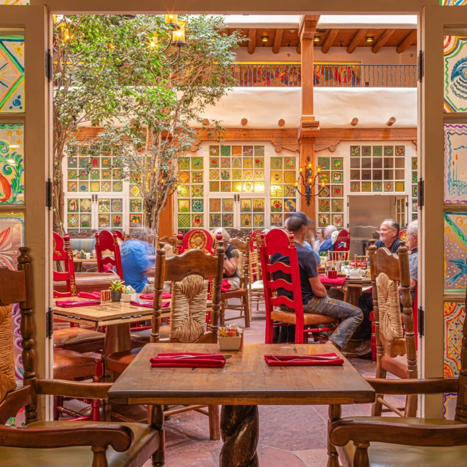 Guests are seated at wooden tables inside a vibrant restaurant, featuring red chairs and colorful tiled windows.