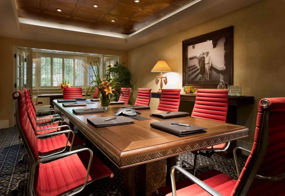A large, professional meeting room features a carved wooden table, high-backed red chairs, and soft lighting.